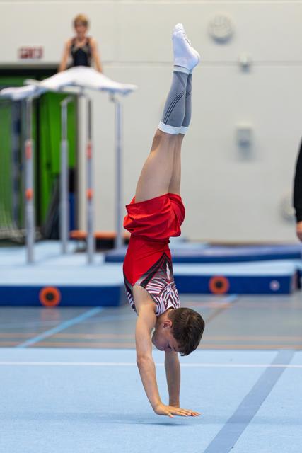 Young athlete performs a handstand on training floor, wearing red shirt and patterned shorts, legs pointed skyward