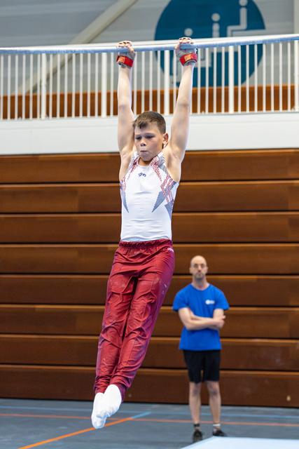 Young athlete hangs from horizontal bar with arms fully extended, wearing white and burgundy uniform in indoor sports hall