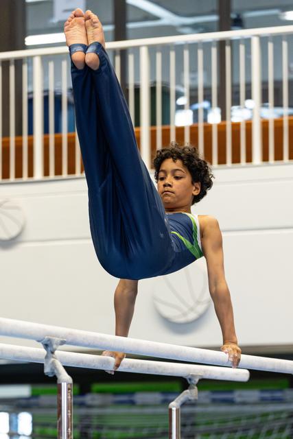 Young gymnast hangs from parallel bars with legs extended upward in pike position, demonstrating strength and concentration