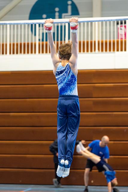 Young athlete in blue leotard hangs from high bar with arms extended overhead during indoor training session
