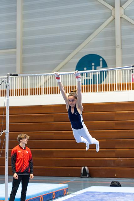 Young gymnast hanging with arms extended on high bar during training, coach observing below in indoor sports hall