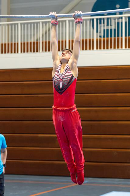 Young athlete in red uniform hangs from high bar with arms fully extended, looking up during training in indoor gymnasium
