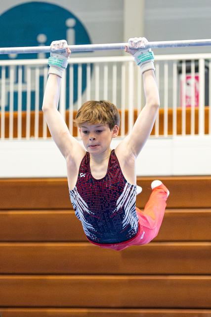 Young gymnast hanging from horizontal bar with arms extended overhead, wearing sparkly leotard and pink tights in training facility