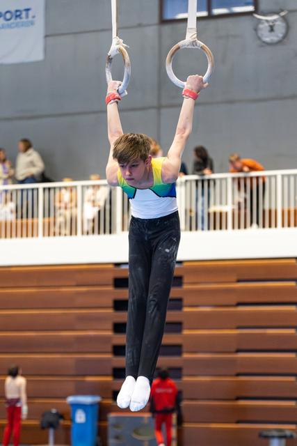Young athlete performs a hang position on still rings, arms extended overhead, wearing a colorful leotard at an indoor venue