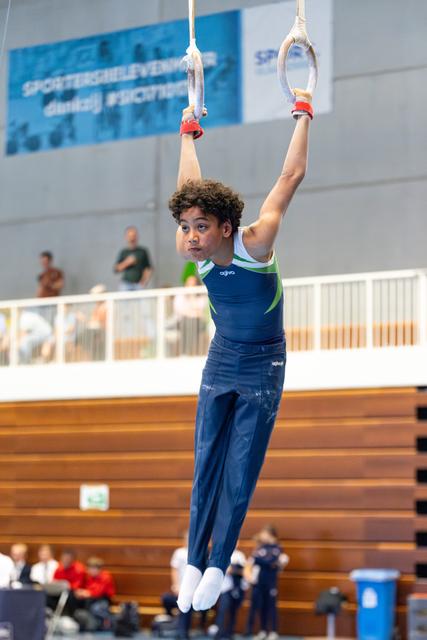 Young athlete hanging from rings with focused expression during routine, wooden floor and spectators visible in background