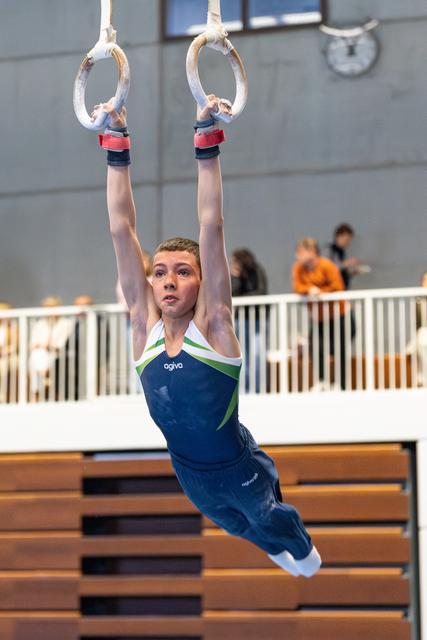 Young athlete hanging from still rings with arms extended overhead during routine, wearing navy blue leotard in indoor venue