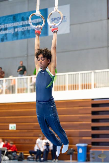 Young athlete grips still rings during routine, body fully extended with focused expression in indoor sports hall