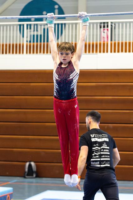 Young athlete hangs from horizontal bar with focused expression while coach spots from below in indoor training facility