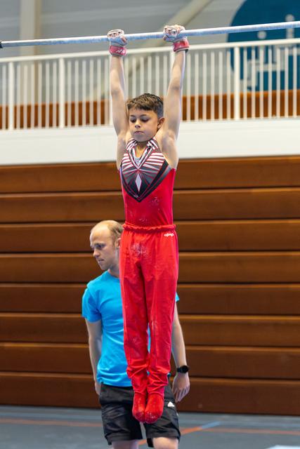 Young athlete in red outfit hanging from horizontal bar with arms extended while coach in blue shirt spots from below