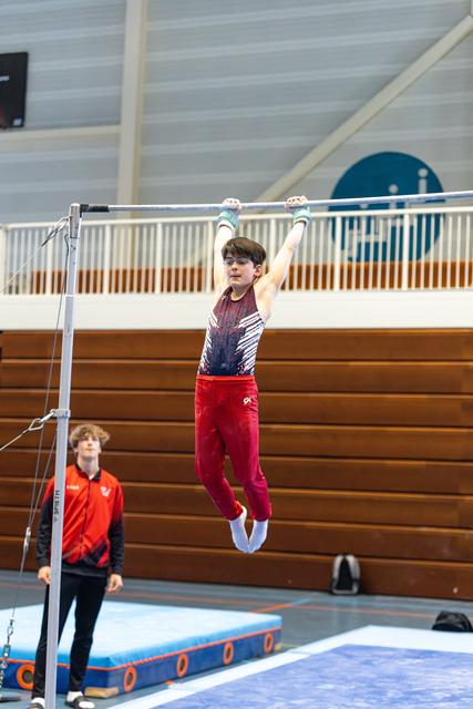 Young athlete hanging from horizontal bar with arms extended, wearing red pants and gradient leotard, coach watching below