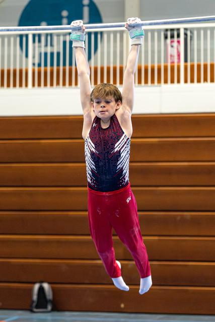 Young gymnast hangs from horizontal bar with arms extended overhead in red and black leotard during training session