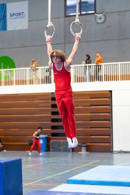 Young athlete in red uniform hanging from rings mid-air, displaying strength and focus during routine at indoor sports facility
