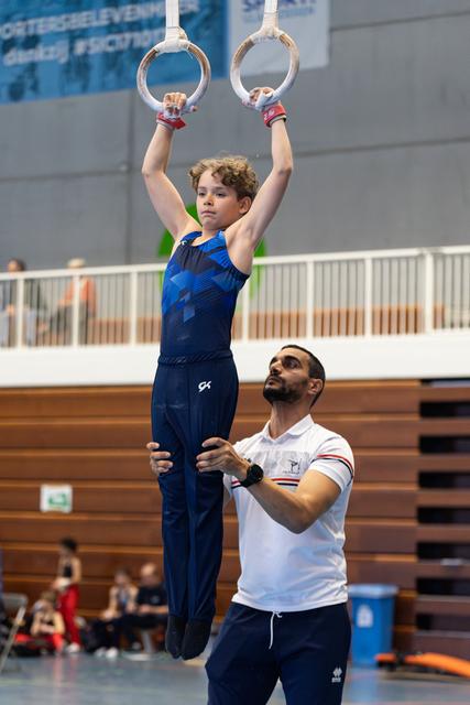 Young gymnast hanging from rings with focused expression while coach stands below providing support during training