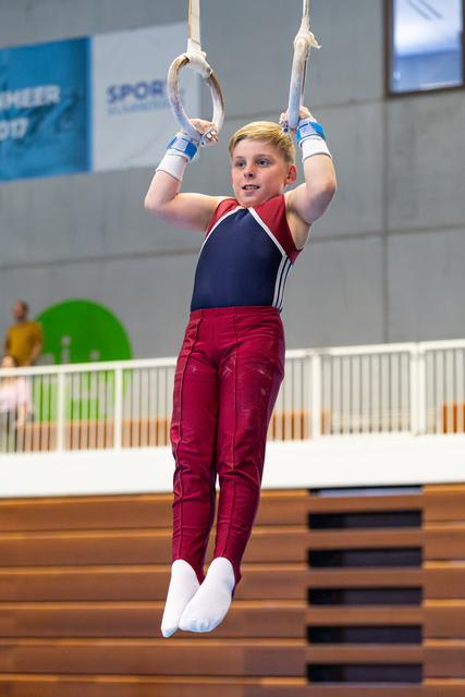Young athlete hanging from rings in red and navy uniform, displaying focus during training at indoor sports facility