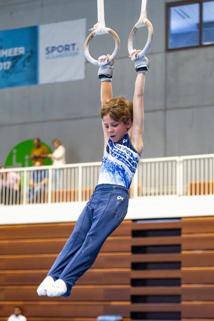 Young gymnast hanging from rings with focused expression during training, wearing blue and white outfit in sports hall