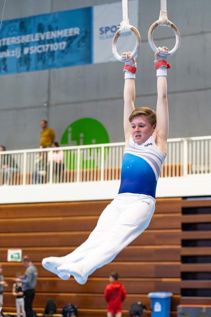 Young gymnast hanging from still rings in white and blue uniform, demonstrating strength hold in indoor sports hall
