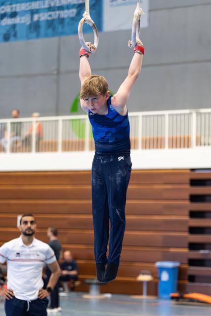 Young athlete with wrists wrapped hangs from rings during training, focused expression, coach observing from below in gymnasium
