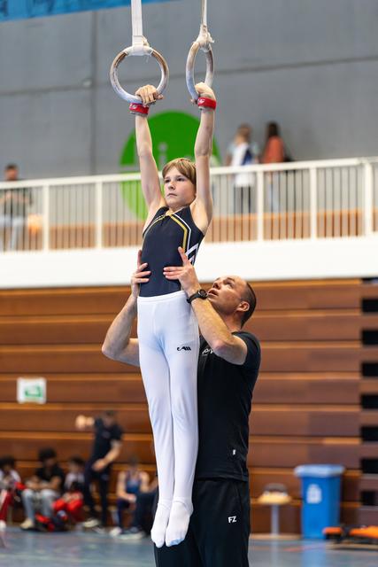 Young gymnast hangs from rings with focused expression while coach provides support and guidance from below in indoor venue