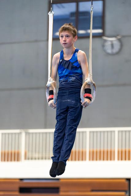 Young athlete in blue uniform hangs from still rings, showing focused expression during training session in gymnasium