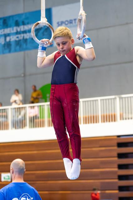 Young athlete in maroon and navy uniform hanging from still rings with focused expression during indoor training
