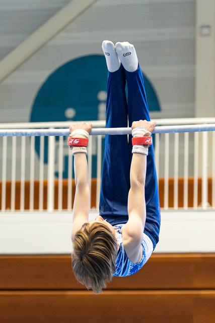 Young athlete hanging upside down from horizontal bar, legs extended upward, wearing blue uniform and white grips