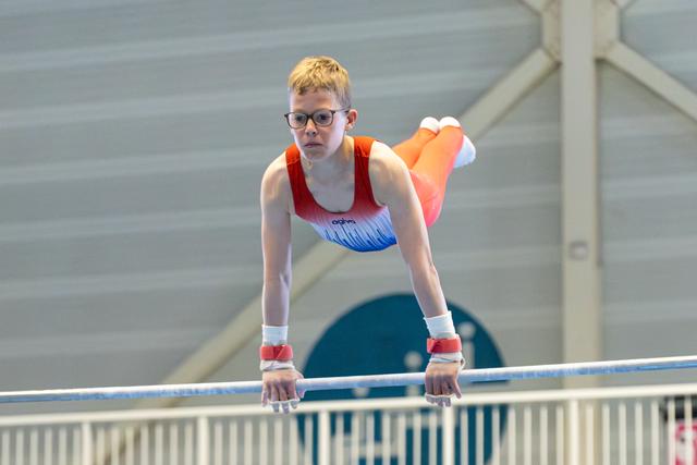 Young gymnast with blonde hair and glasses performs on horizontal bar with intense concentration, wearing red and blue leotard