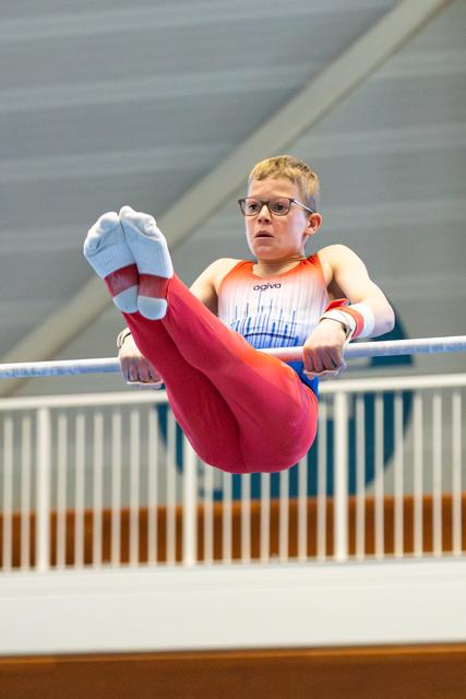 Young athlete with glasses performs horizontal bar routine in red and blue uniform, showing focused concentration