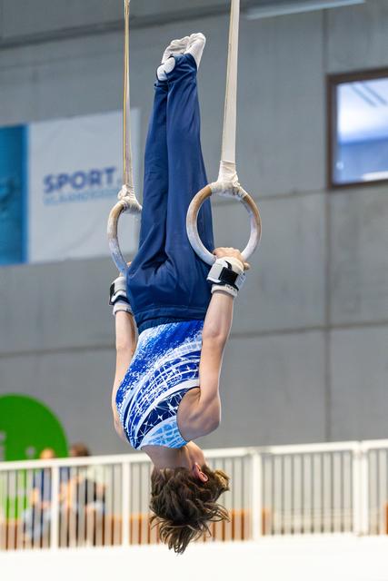 Young athlete performing an inverted hang on still rings, legs extended upward, displaying strength and control in indoor gym