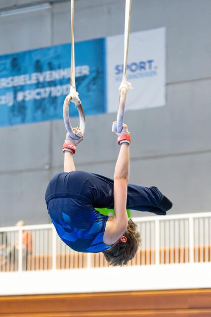 Athlete performing inverted hang on rings, wearing blue outfit with red wrist guards, demonstrating strength during routine