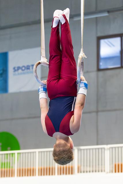 Young athlete performs an inverted hang on rings during routine, displaying strength and control in maroon and navy uniform