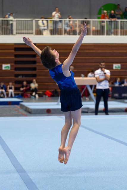 Young athlete mid-jump with arms raised skyward during floor routine, displaying graceful form in indoor sports hall