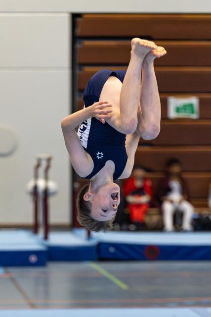 Young athlete performs a tucked backflip mid-air during a gymnastics competition in an indoor sports hall