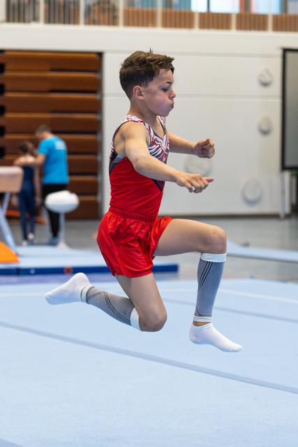 Young athlete jumping mid-air during floor exercise routine in gymnasium, wearing red shorts and striped leotard