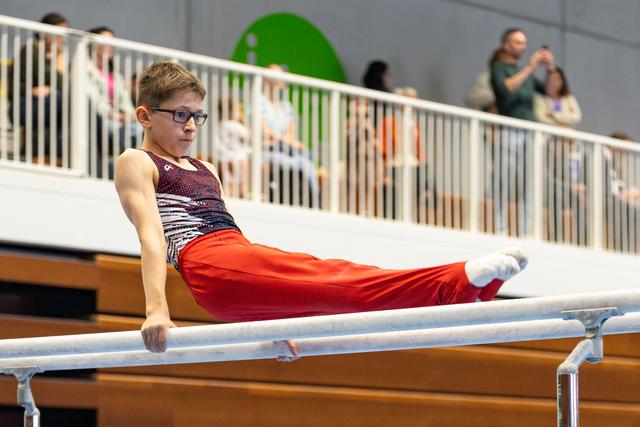 Young athlete with glasses performs parallel bars routine in intense concentration, wearing burgundy leotard and red pants
