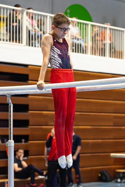 Young gymnast in red pants and patterned leotard performing on parallel bars with focused expression, spectators watching