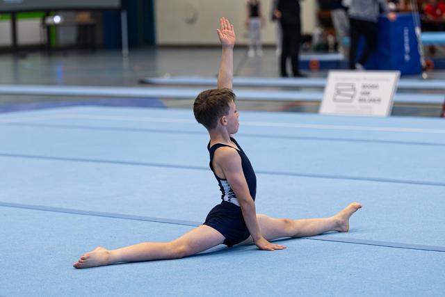Young gymnast performs a split with one arm raised during floor exercise at indoor sports competition