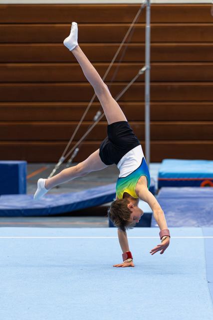 Young gymnast executing a one-handed handstand on floor exercise mat, demonstrating strength and balance in training facility