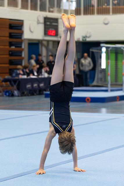Gymnast executing a vertical handstand on floor exercise mat, legs extended upward, with spectators in background