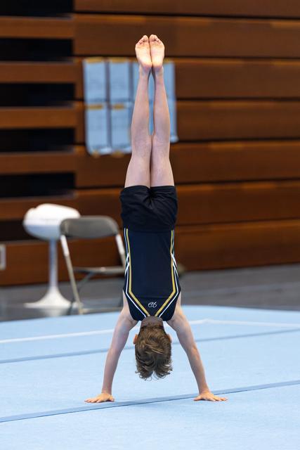 Athlete performs a vertical handstand with pointed toes on blue mat in gymnasium with bleachers in background