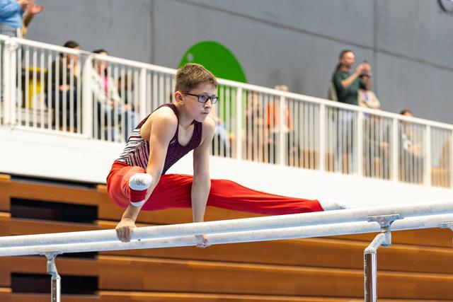 Young athlete in burgundy leotard and red pants performs on parallel bars while spectators watch from balcony above