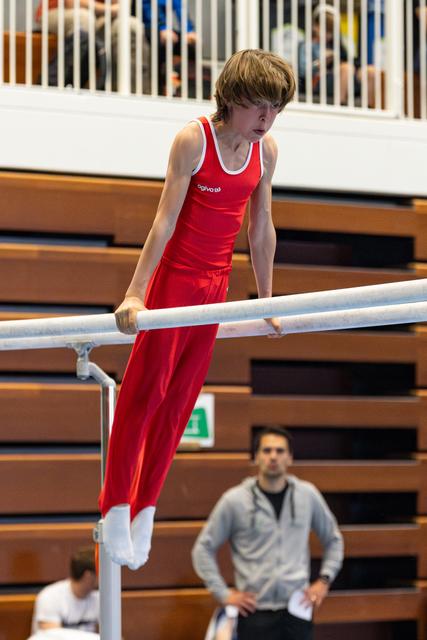 Young gymnast in red uniform demonstrates strength on parallel bars while coach observes from below in competition venue