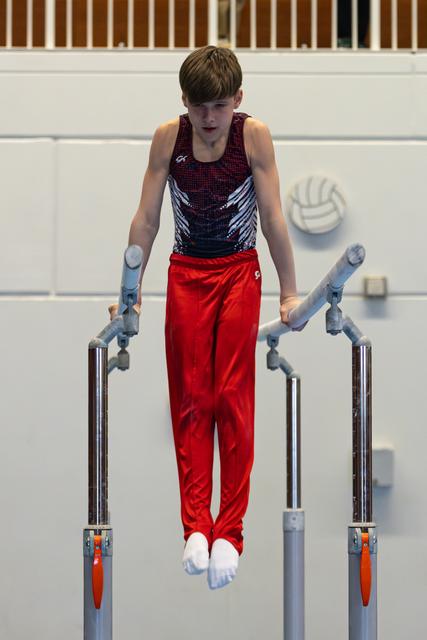 Young athlete performing on parallel bars with focused expression, wearing red pants and patterned leotard in gymnasium