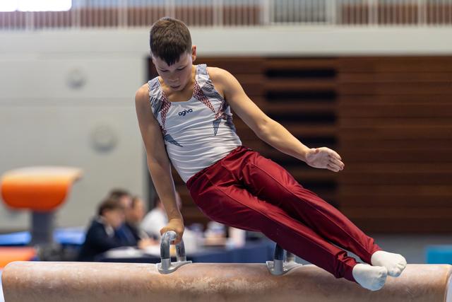 Young athlete performing a pommel horse routine with focused concentration, wearing white and red uniform at indoor competition