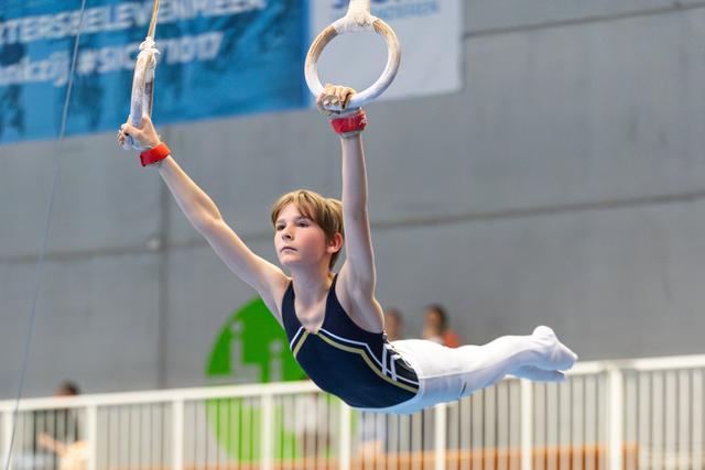 Young gymnast performs still ring hold with focused expression, arms extended during competitive routine at indoor facility