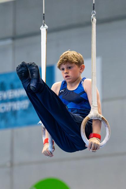 Young athlete demonstrating a tuck position on the rings, focused expression, training facility with pool in background