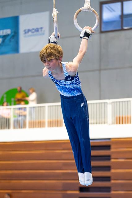 Young athlete performing on rings with focused expression, wearing blue gradient leotard in indoor gymnasium