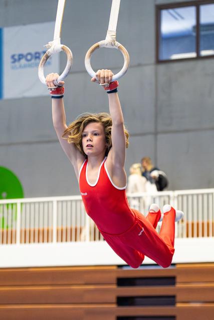 Young gymnast in red leotard performing on rings with focused expression, suspended mid-routine at indoor sports facility
