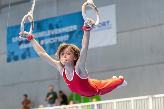 Young athlete in red leotard performs on rings with focused expression, arms raised overhead during routine at indoor venue