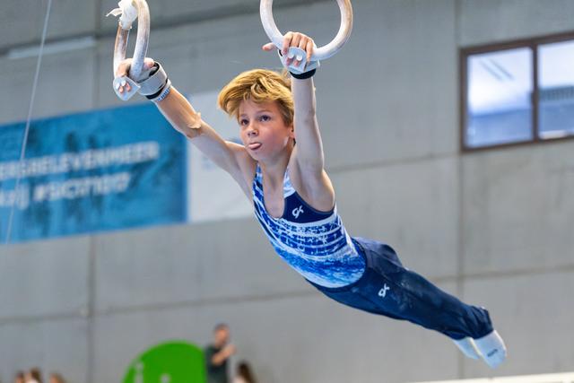 Young athlete suspended mid-air on rings, arms extended upward in perfect form during competitive routine at indoor facility