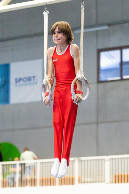 Young athlete suspended on still rings in red uniform, showing focused concentration during routine at indoor training facility